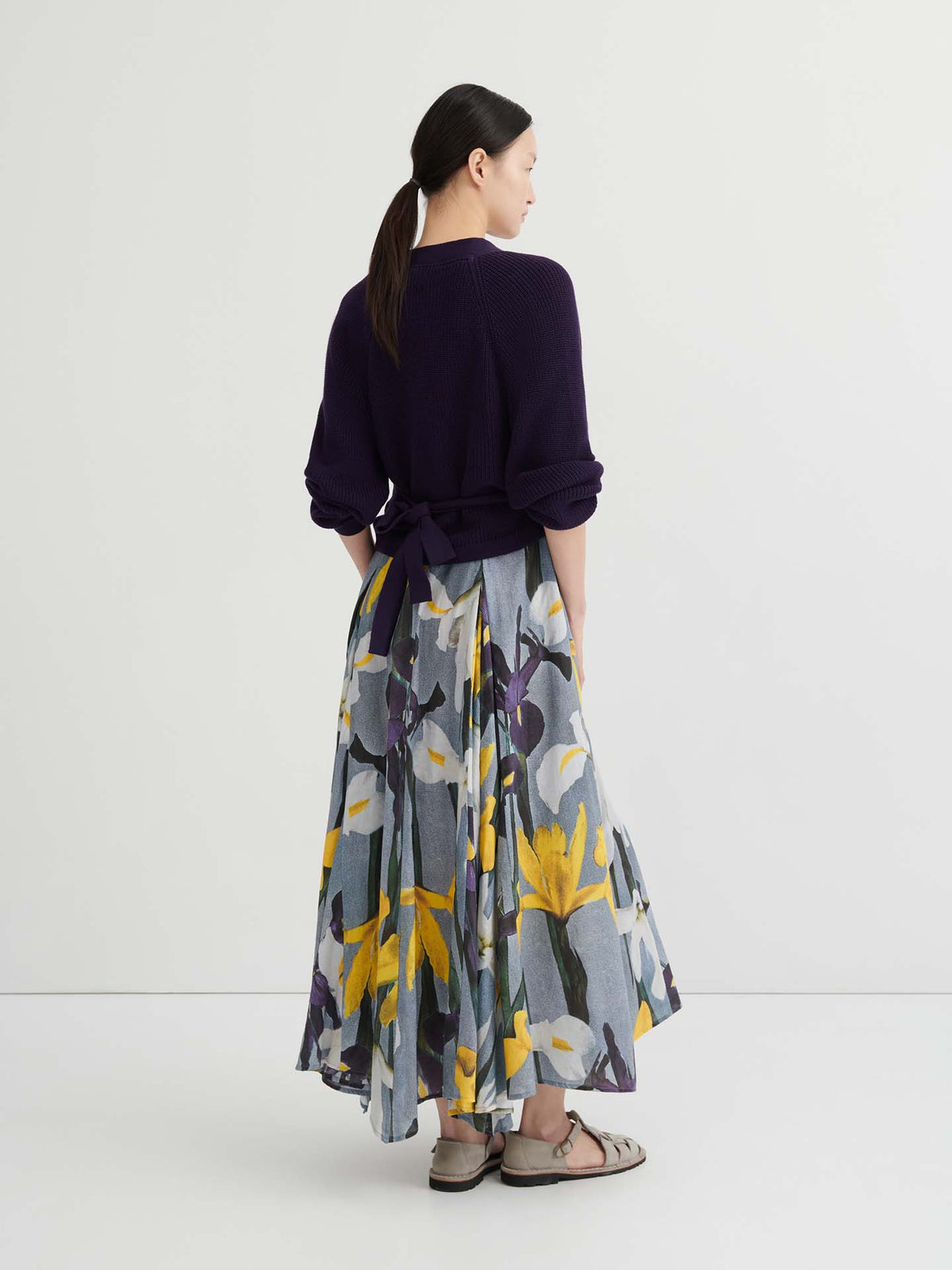 A woman stands with her back to the camera, wearing a relaxed fit Composure Cardigan in Dahlia by Kowtow, a floral print skirt featuring yellow and white flowers, and sandals.