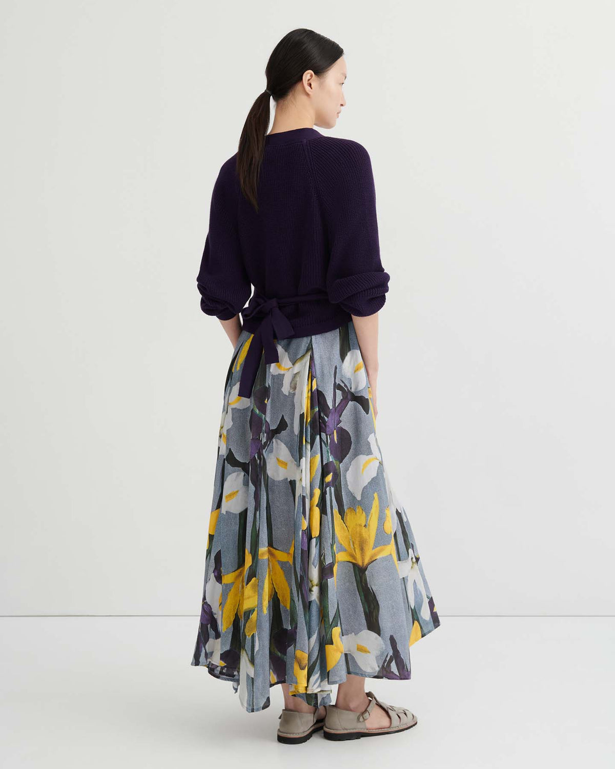 A woman stands with her back to the camera, wearing a relaxed fit Composure Cardigan in Dahlia by Kowtow, a floral print skirt featuring yellow and white flowers, and sandals.