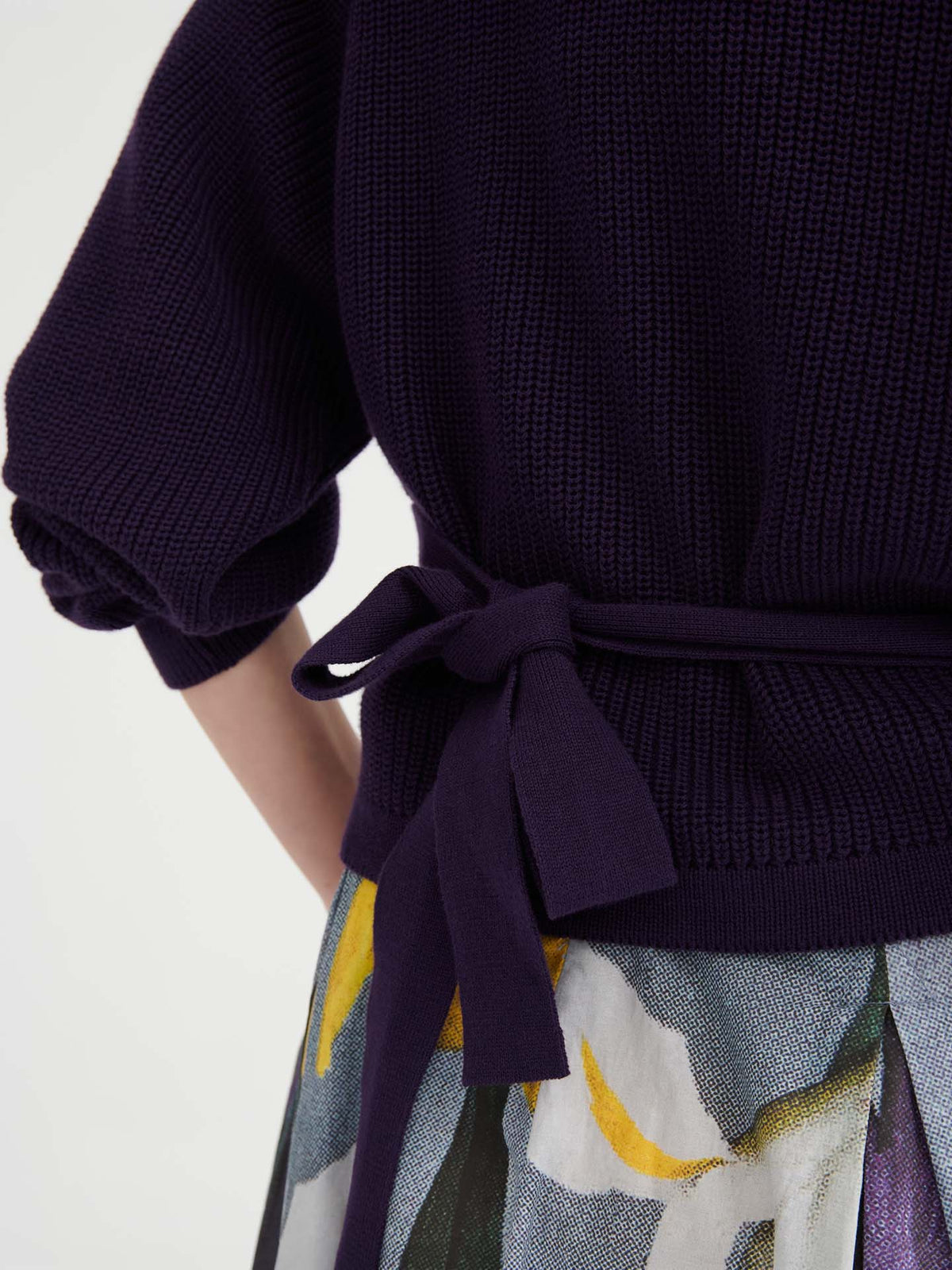 Close-up of a person wearing the Composure Cardigan – Dahlia in a dark knit with a tied waist belt and a skirt featuring a colorful abstract pattern, highlighting the relaxed fit and effortless style.