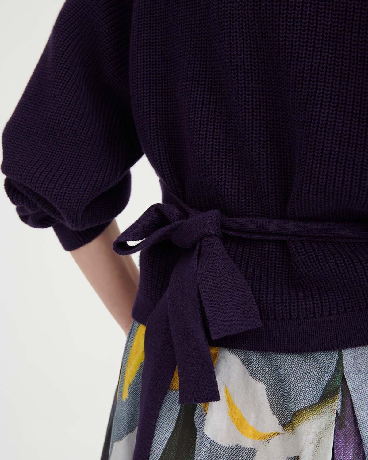 Close-up of a person wearing the Composure Cardigan – Dahlia in a dark knit with a tied waist belt and a skirt featuring a colorful abstract pattern, highlighting the relaxed fit and effortless style.