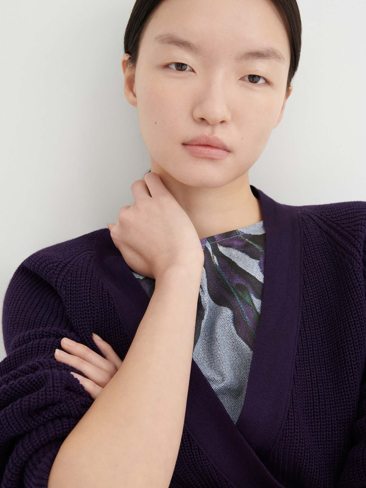 A woman with straight dark hair is wearing the Composure Cardigan – Dahlia, featuring a relaxed fit in deep purple, layered over a printed top. She stands against a plain white background with a neutral expression.