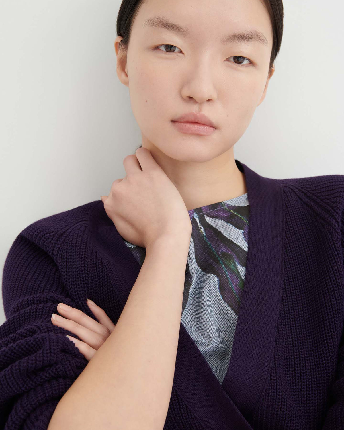 A woman with straight dark hair is wearing the Composure Cardigan – Dahlia, featuring a relaxed fit in deep purple, layered over a printed top. She stands against a plain white background with a neutral expression.