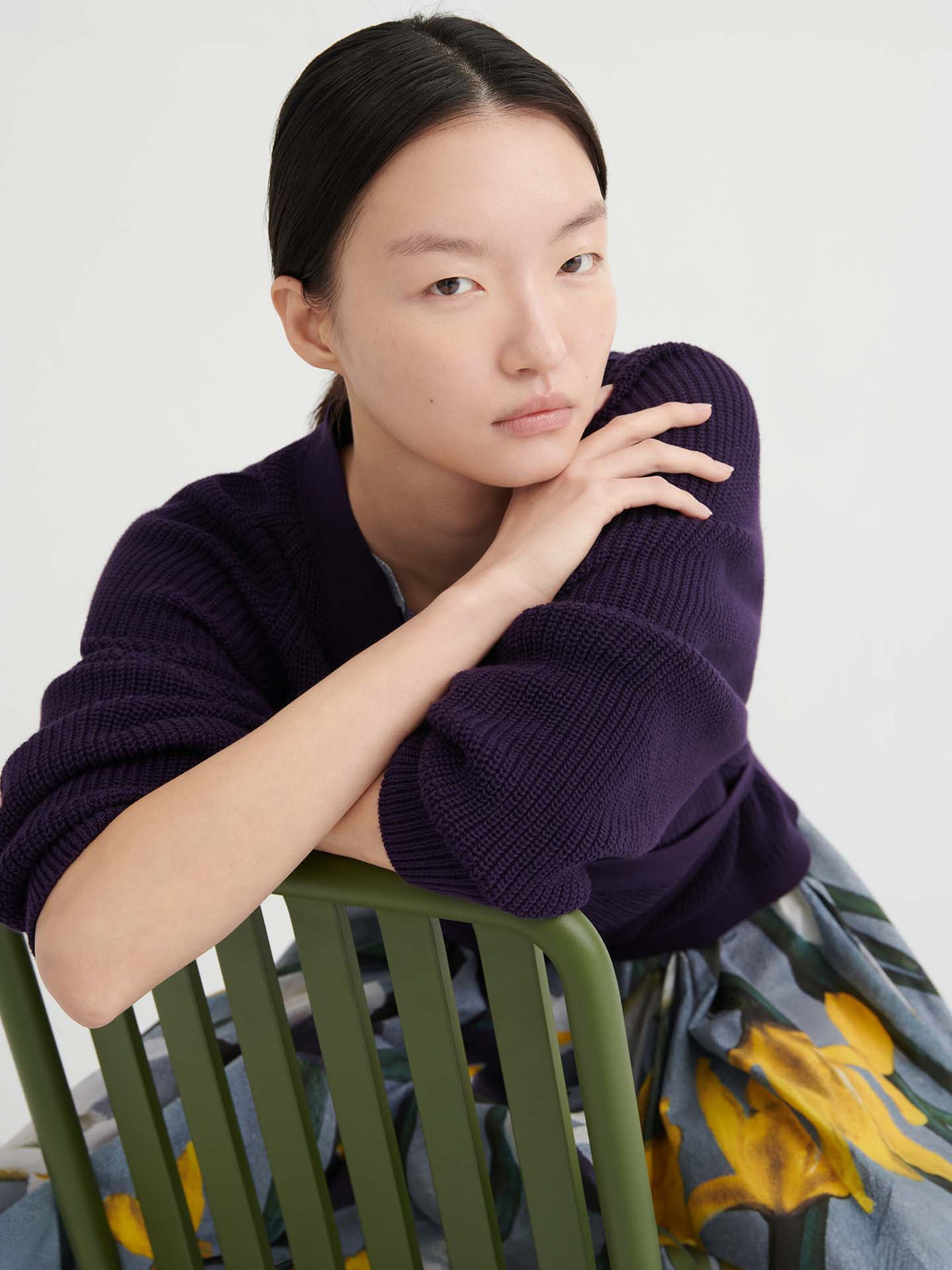 Person with straight black hair, wearing a relaxed fit Composure Cardigan – Dahlia from Kowtow and a floral skirt, sits on a green chair, resting their arms on the chair back and looking at the camera with a neutral expression.