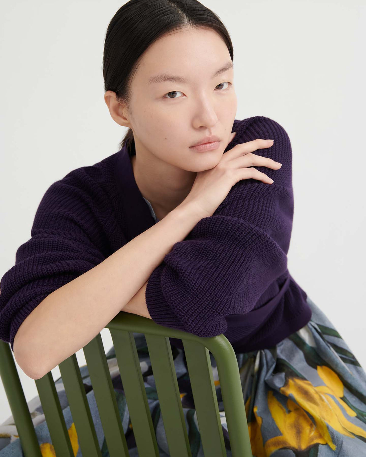 Person with straight black hair, wearing a relaxed fit Composure Cardigan – Dahlia from Kowtow and a floral skirt, sits on a green chair, resting their arms on the chair back and looking at the camera with a neutral expression.