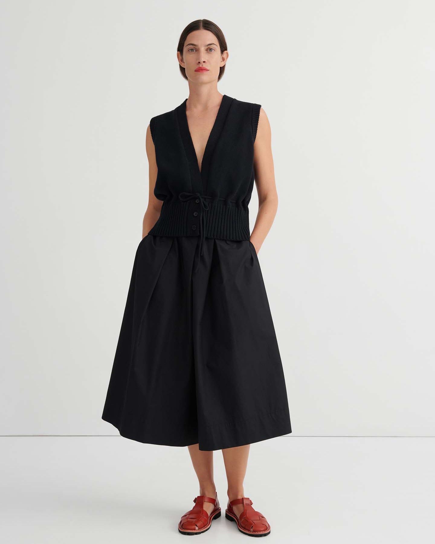 A woman stands against a plain background wearing the Culottes – Black, featuring a relaxed fit, wide-leg silhouette that sits at the natural waist. She pairs them with a sleeveless black vest and red flat shoes, her hands resting casually in her pockets.