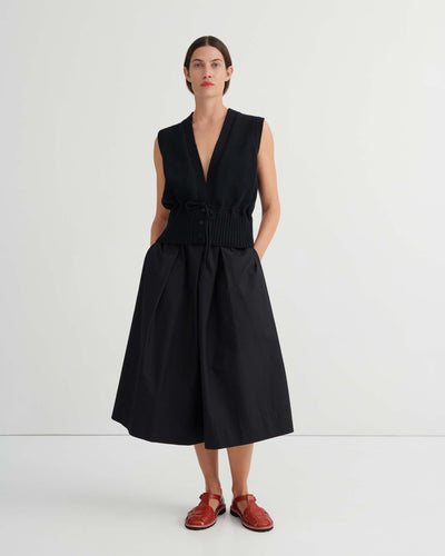 A woman stands against a plain background wearing the Culottes – Black, featuring a relaxed fit, wide-leg silhouette that sits at the natural waist. She pairs them with a sleeveless black vest and red flat shoes, her hands resting casually in her pockets.