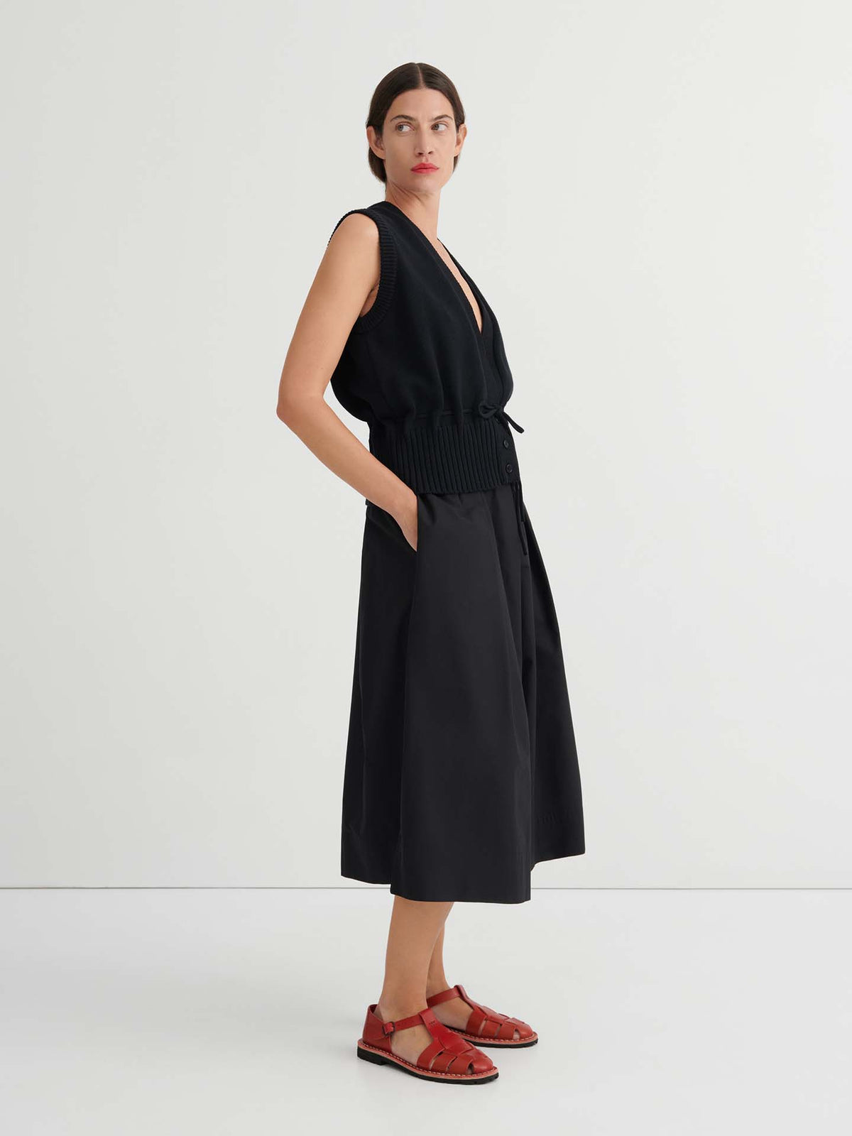 A woman stands against a plain white background, wearing black Culottes – Black with a natural waist and relaxed fit, paired with a sleeveless black top and red sandals, her hands in her pockets.
