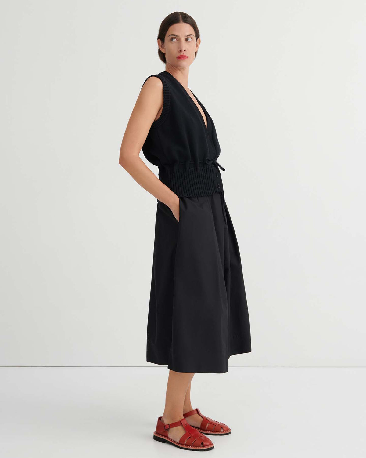 A woman stands against a plain white background, wearing black Culottes – Black with a natural waist and relaxed fit, paired with a sleeveless black top and red sandals, her hands in her pockets.