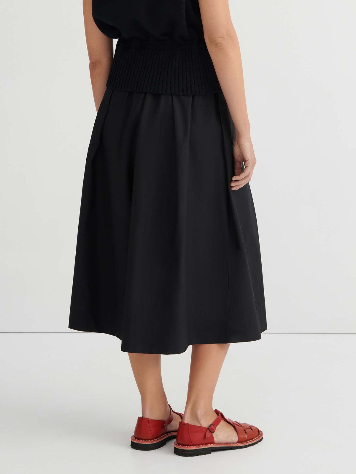 A person is seen from behind, standing against a plain light background and wearing the Culottes – Black with a natural waist, styled with a black top and red closed-toe sandals.