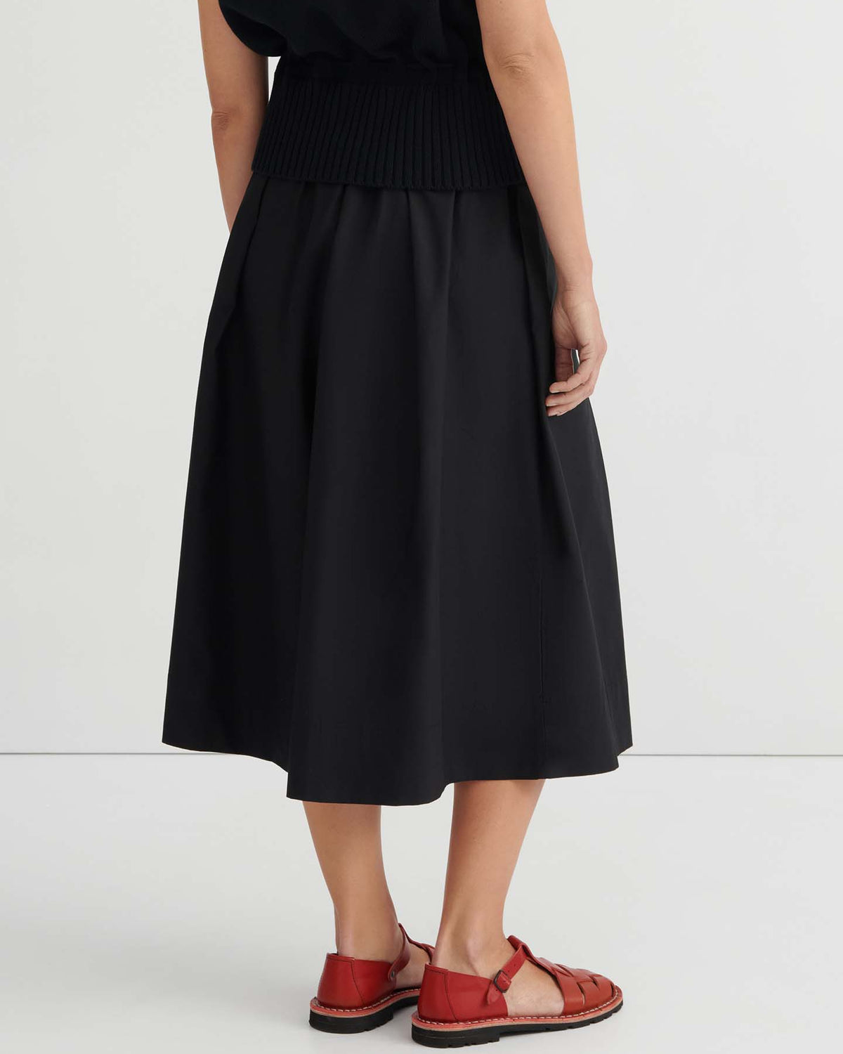 A person is seen from behind, standing against a plain light background and wearing the Culottes – Black with a natural waist, styled with a black top and red closed-toe sandals.