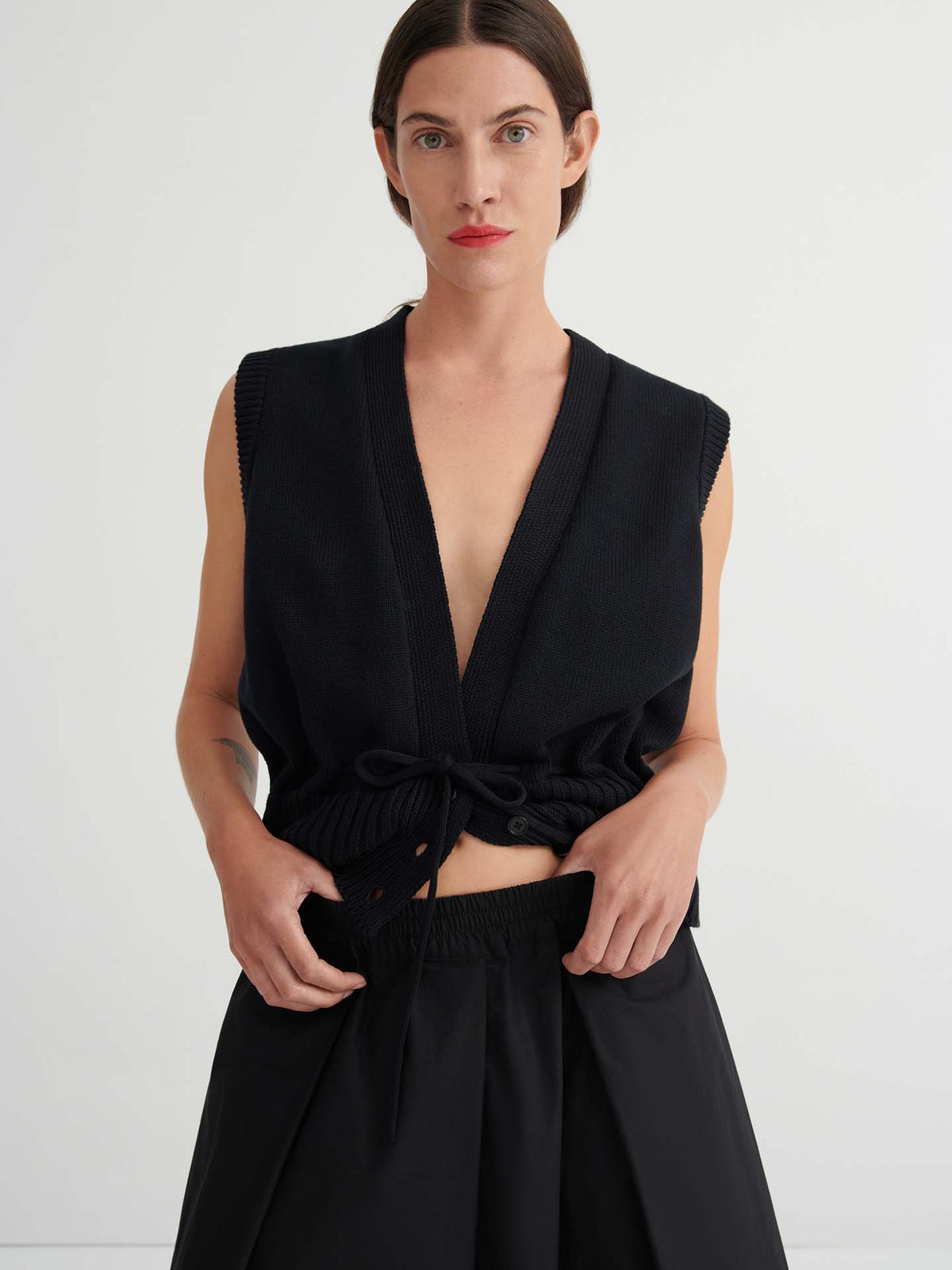A woman with straight brown hair wears a sleeveless black V-neck top with drawstring and Culottes – Black with a natural waist, standing against a plain light background.