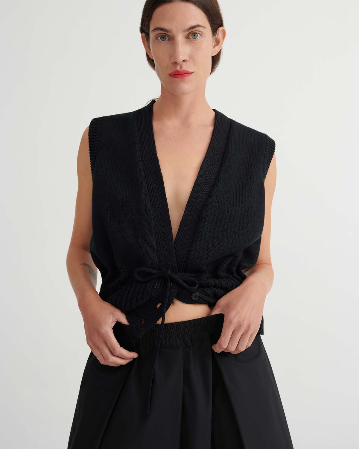 A woman with straight brown hair wears a sleeveless black V-neck top with drawstring and Culottes – Black with a natural waist, standing against a plain light background.