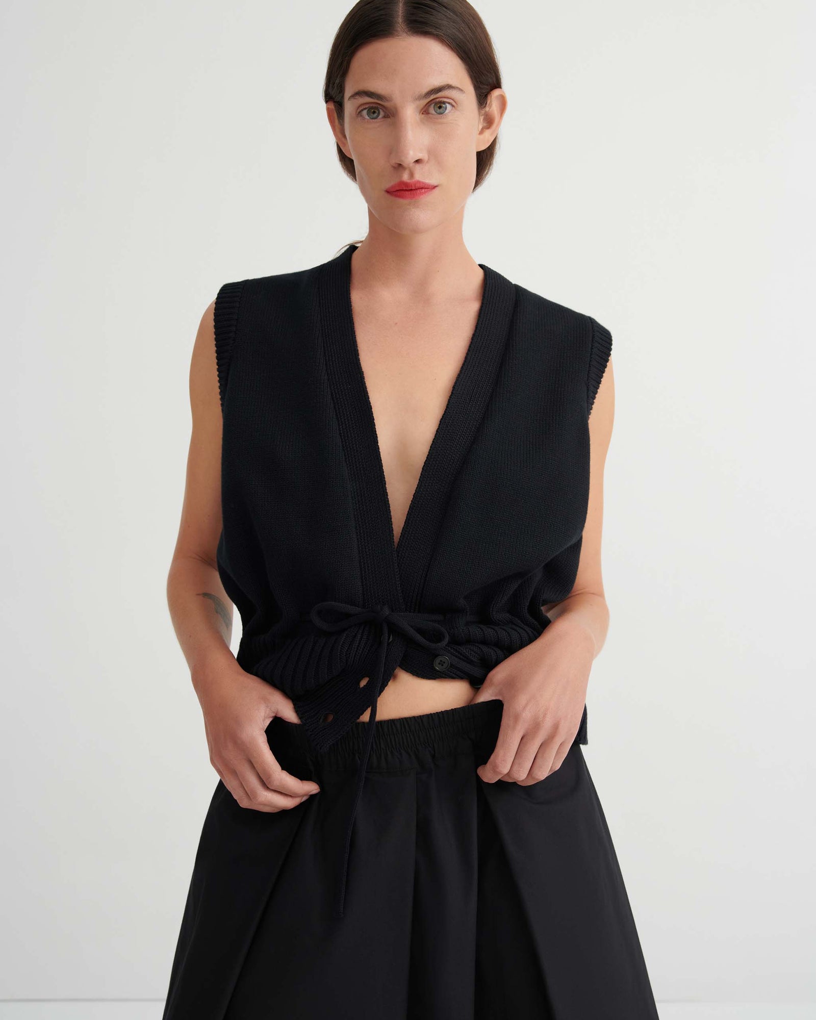 A woman stands against a plain background wearing the Culottes – Black, featuring a relaxed fit, wide-leg silhouette that sits at the natural waist. She pairs them with a sleeveless black vest and red flat shoes, her hands resting casually in her pockets.