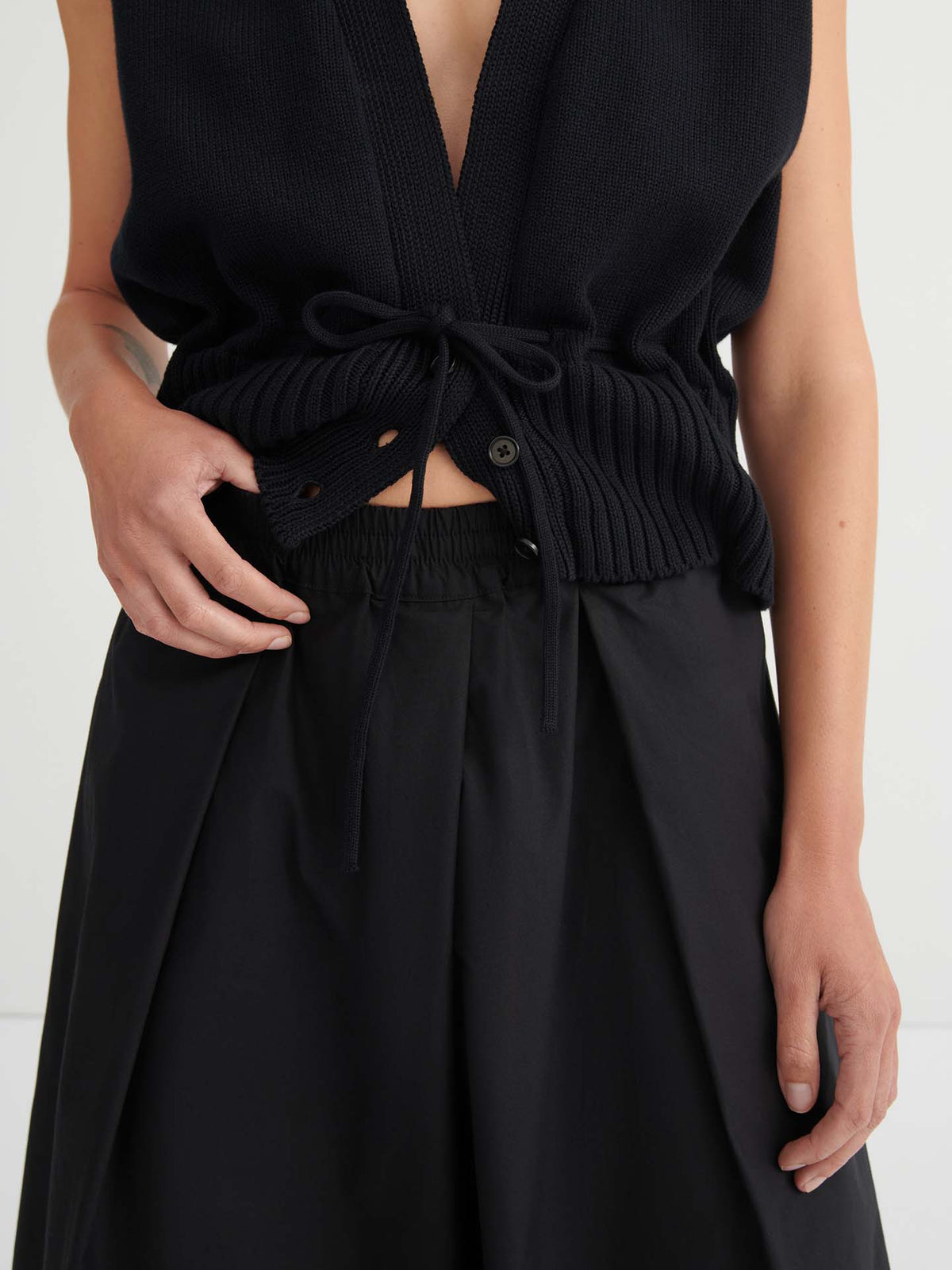 A person is shown from the torso down, wearing a sleeveless black knit top featuring a drawstring and button detail, paired with the Culottes – Black.