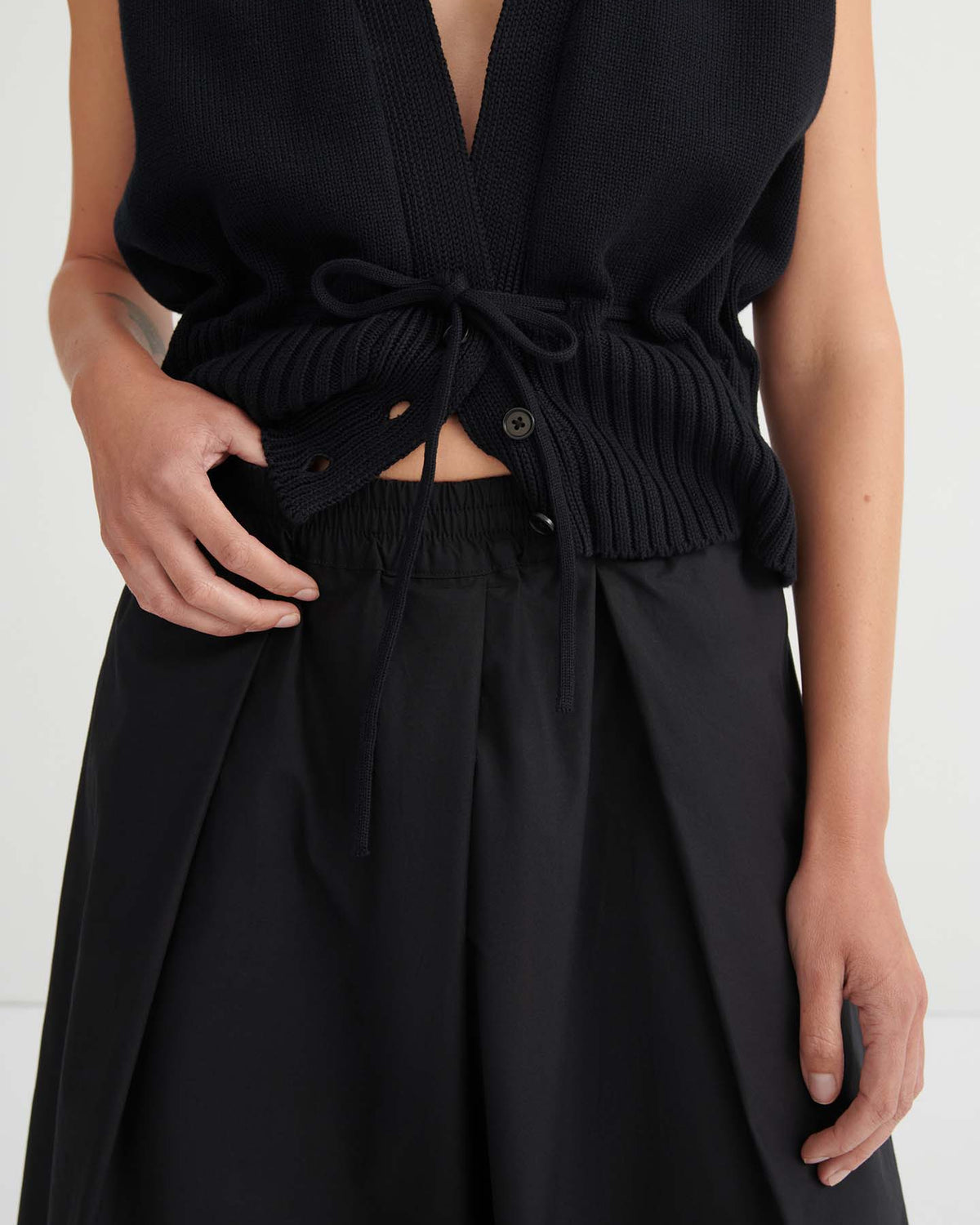 A person is shown from the torso down, wearing a sleeveless black knit top featuring a drawstring and button detail, paired with the Culottes – Black.