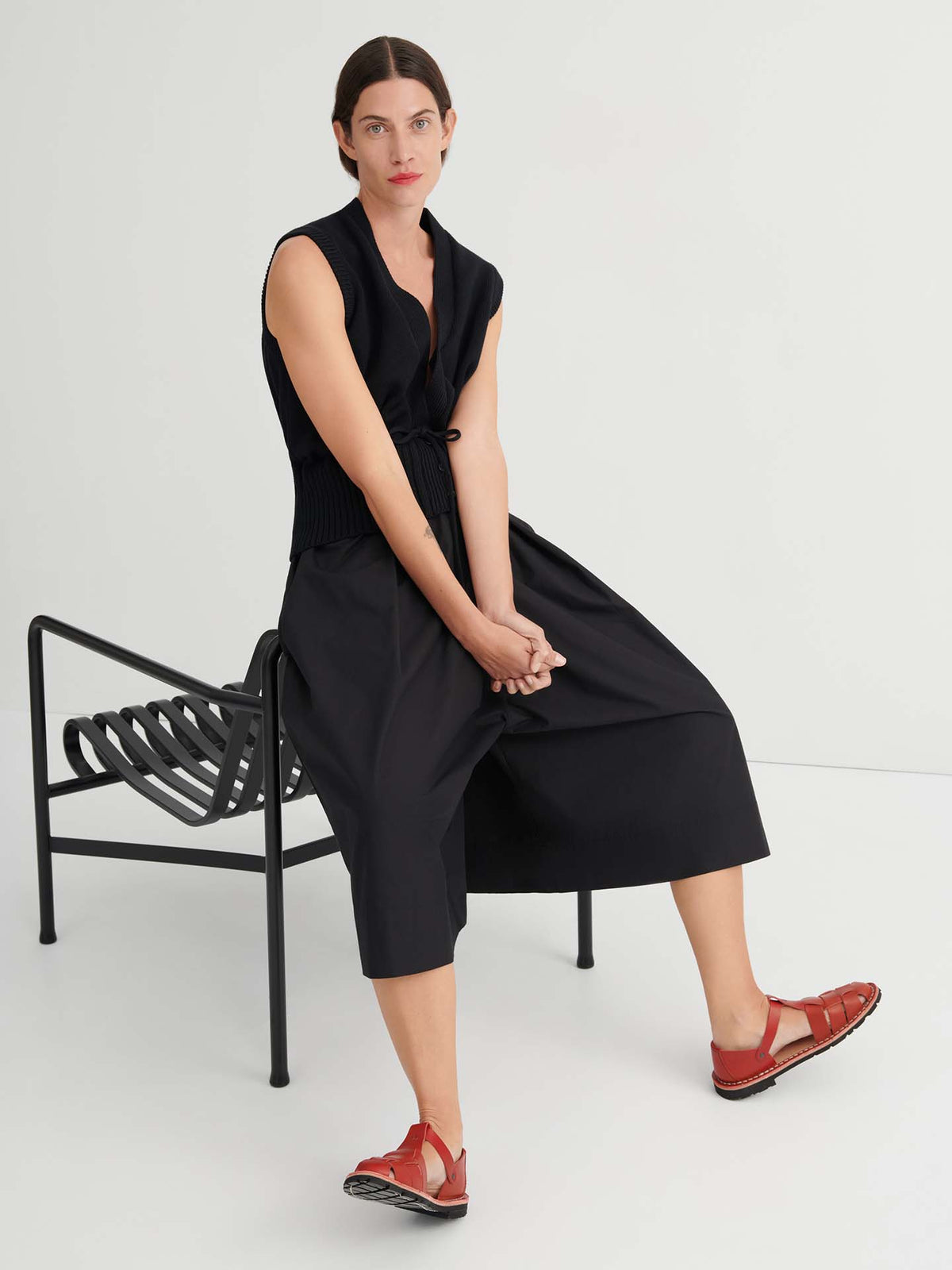 A woman with dark hair sits on a black bench, wearing a sleeveless black top and the Culottes – Black, which feature a relaxed fit and sit at the natural waist, paired with red sandals, against a plain white background.