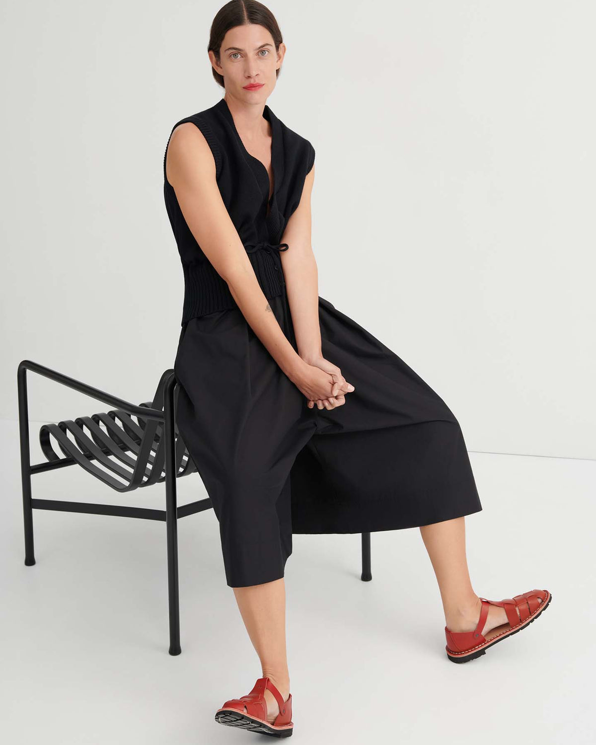 A woman with dark hair sits on a black bench, wearing a sleeveless black top and the Culottes – Black, which feature a relaxed fit and sit at the natural waist, paired with red sandals, against a plain white background.