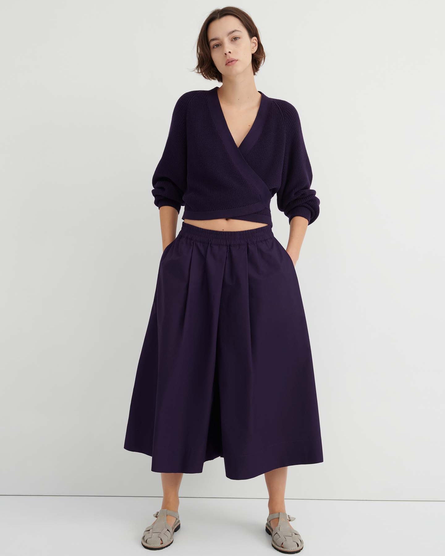A person stands against a white wall wearing a dark purple wrap sweater, the Dahlia culottes—relaxed fit and sitting at the natural waist—with hands in their pockets, paired with gray sandals.