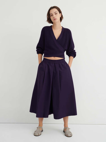 A person stands against a white wall wearing a dark purple wrap sweater, the Dahlia culottes—relaxed fit and sitting at the natural waist—with hands in their pockets, paired with gray sandals.