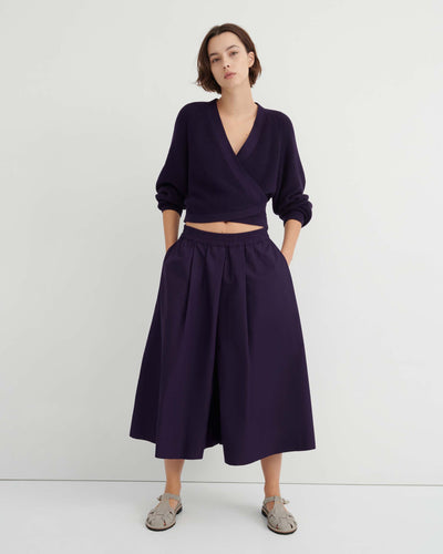 A person stands against a white wall wearing a dark purple wrap sweater, the Dahlia culottes—relaxed fit and sitting at the natural waist—with hands in their pockets, paired with gray sandals.