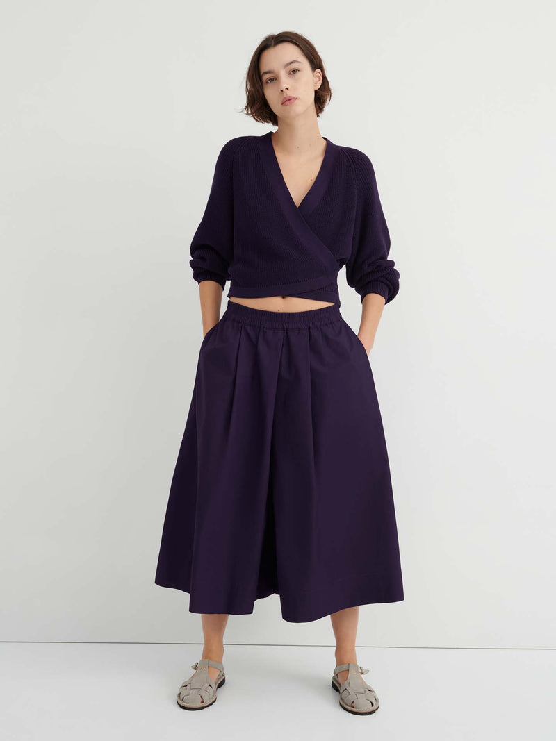 A person stands against a white wall wearing a dark purple wrap sweater, the Dahlia culottes—relaxed fit and sitting at the natural waist—with hands in their pockets, paired with gray sandals.
