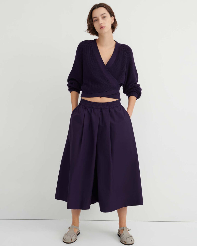 A person stands against a white wall wearing a dark purple wrap sweater, the Dahlia culottes—relaxed fit and sitting at the natural waist—with hands in their pockets, paired with gray sandals.