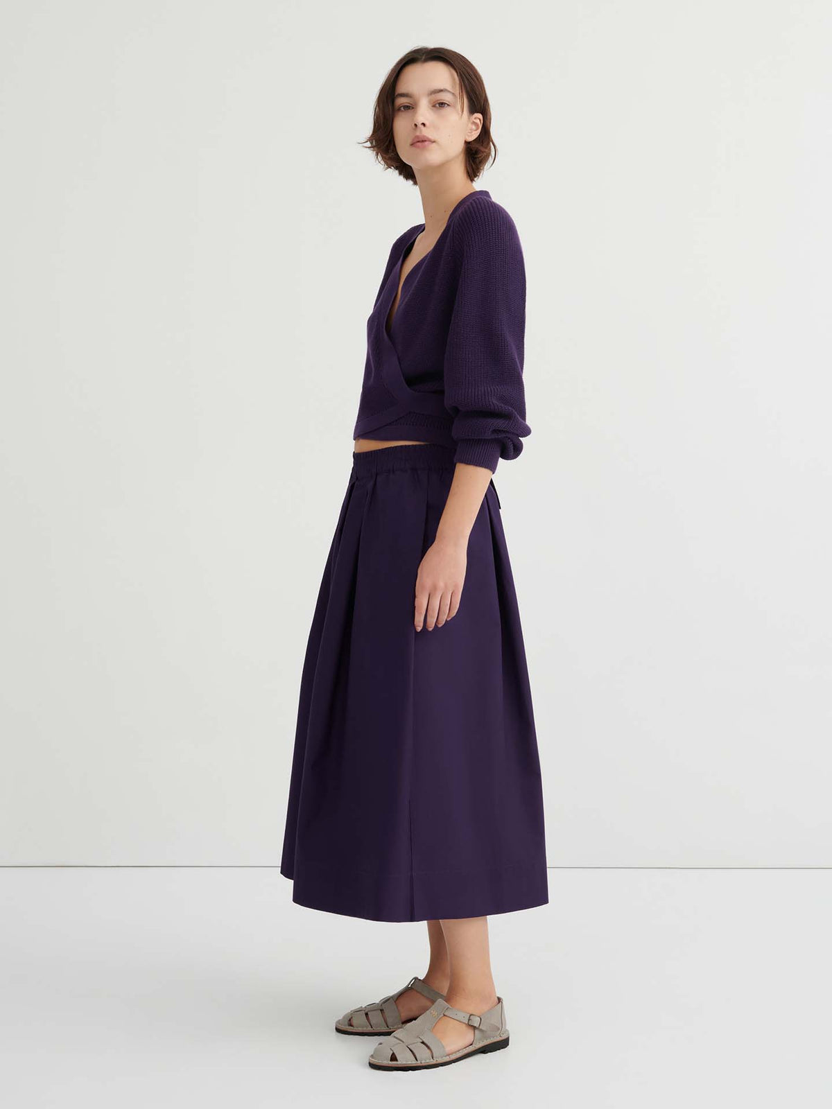 Woman standing sideways wearing a deep purple V-neck sweater with a relaxed fit, matching Culottes – Dahlia, and gray sandals against a plain white background.