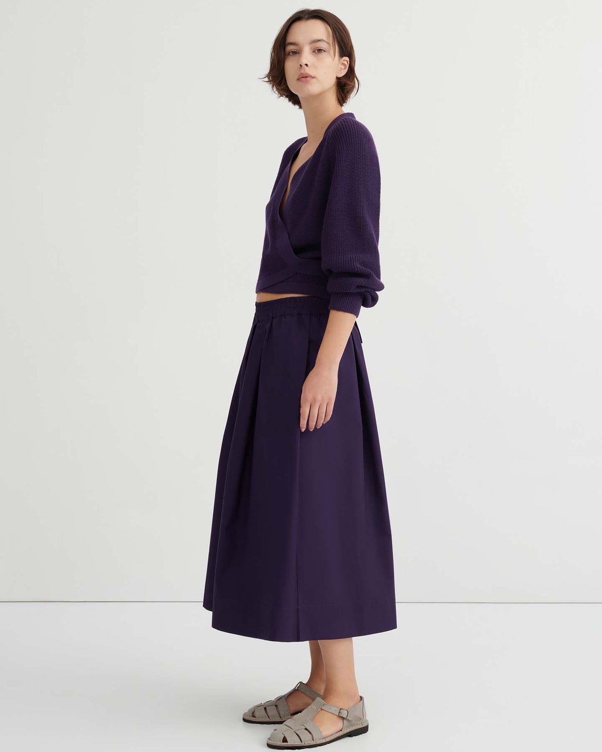 Woman standing sideways wearing a deep purple V-neck sweater with a relaxed fit, matching Culottes – Dahlia, and gray sandals against a plain white background.