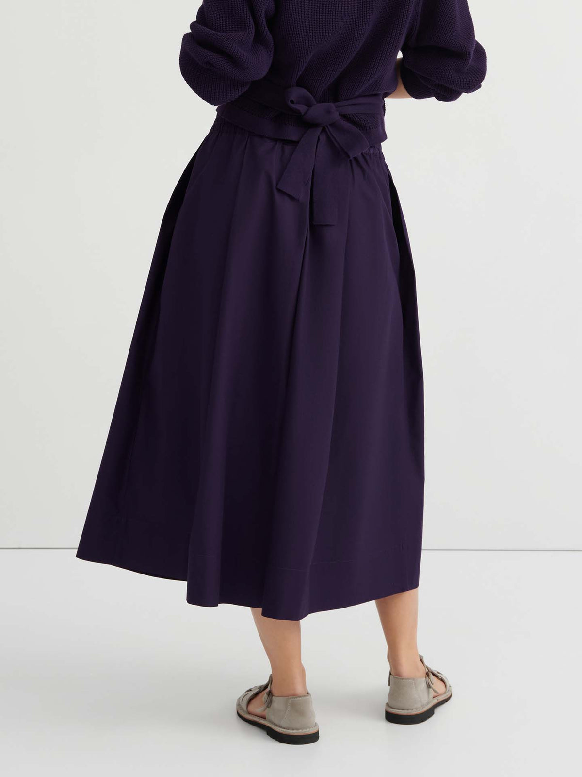 Person standing with back turned, wearing the Culottes – Dahlia in dark purple, featuring a relaxed fit with a natural waist accented by a tied bow, paired with long sleeves and light sandals.
