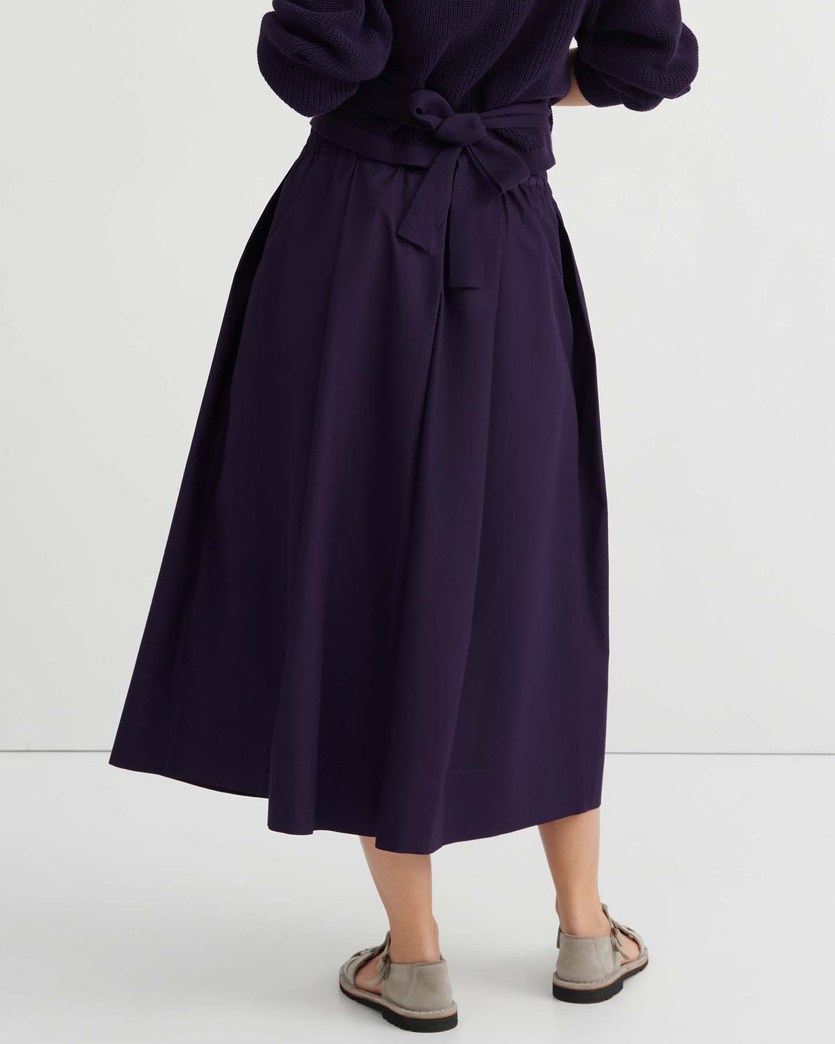 Person standing with back turned, wearing the Culottes – Dahlia in dark purple, featuring a relaxed fit with a natural waist accented by a tied bow, paired with long sleeves and light sandals.
