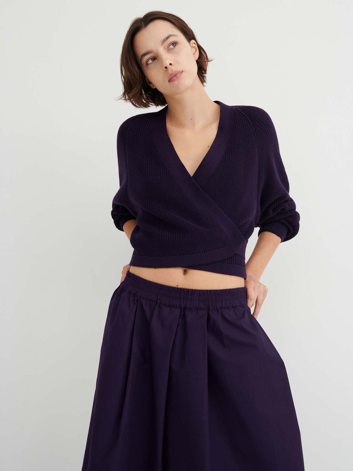 A woman stands against a plain light background, looking slightly to the side with her hands on her hips, wearing relaxed fit Culottes – Dahlia in deep purple and a matching wrap sweater.