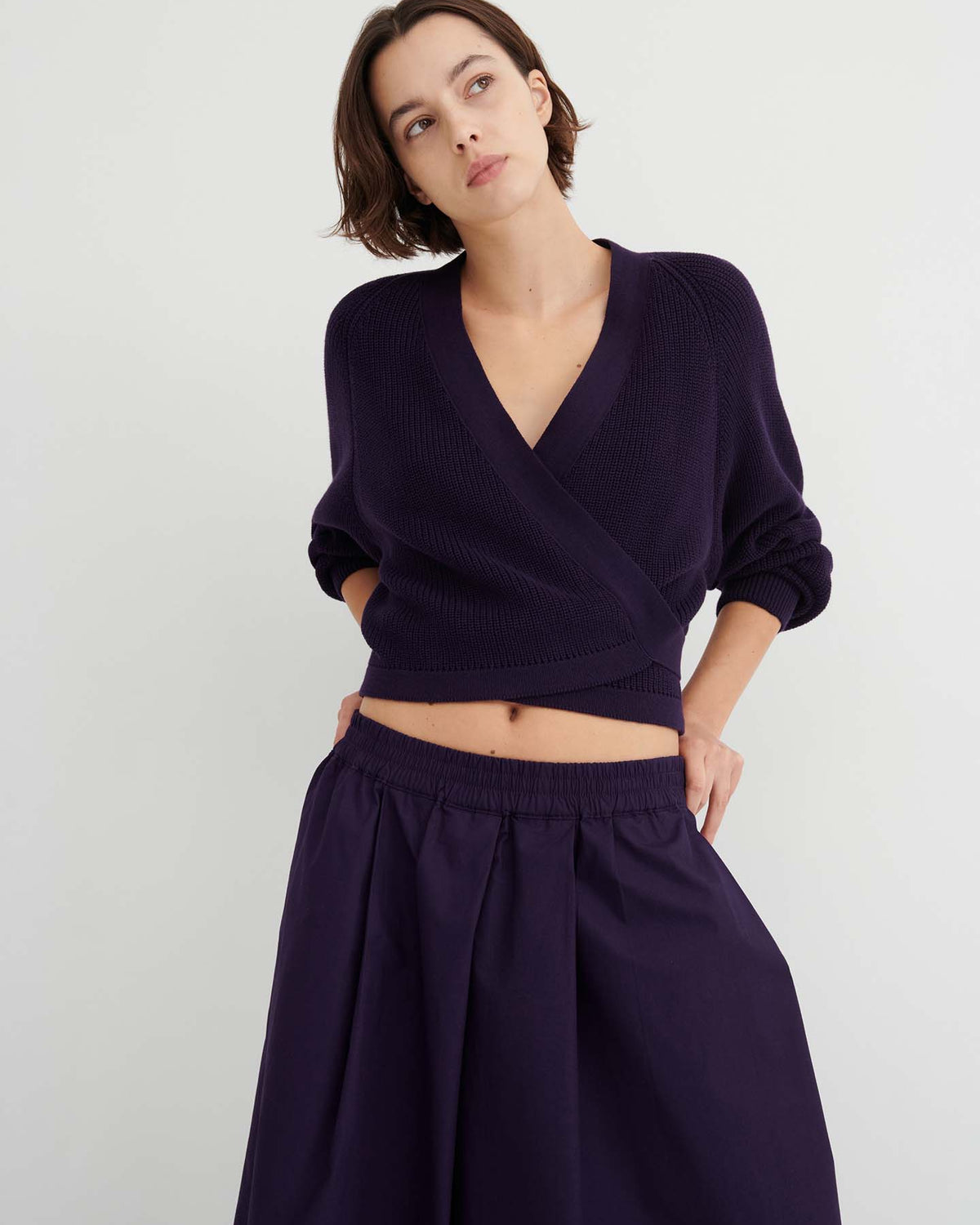 A woman stands against a plain light background, looking slightly to the side with her hands on her hips, wearing relaxed fit Culottes – Dahlia in deep purple and a matching wrap sweater.