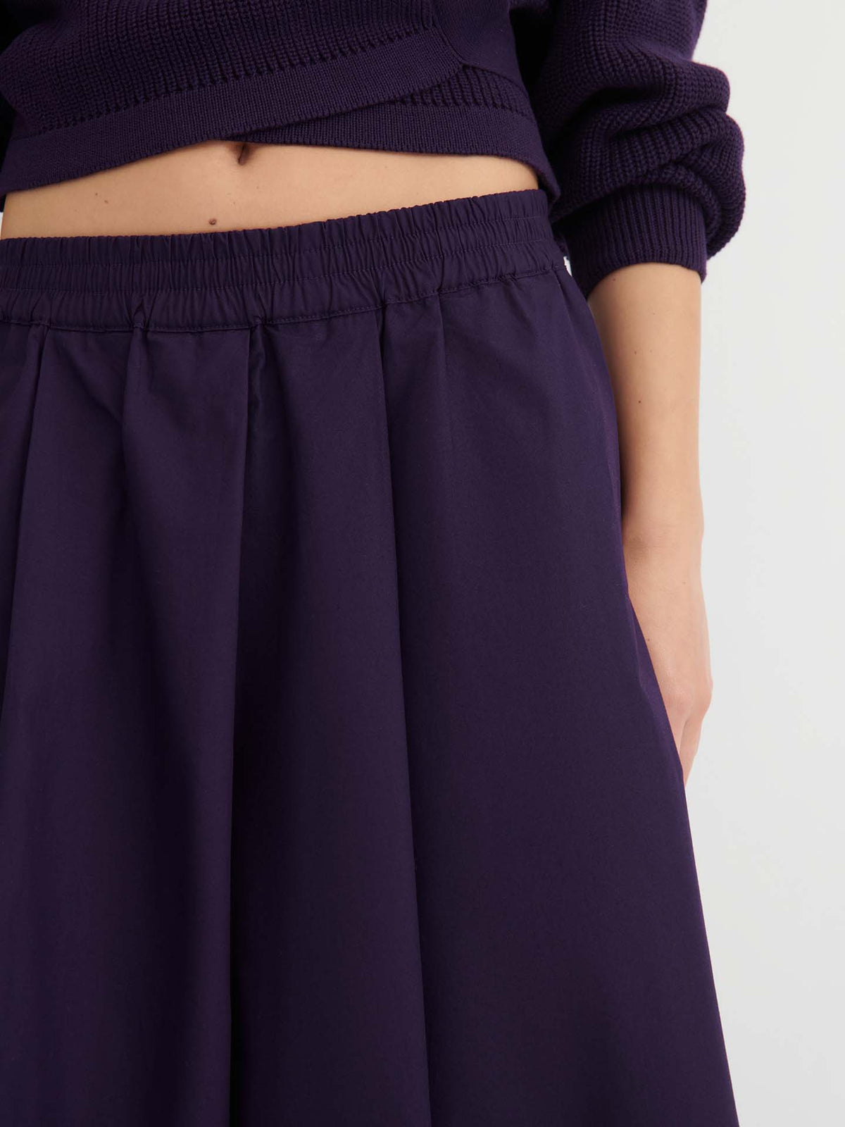 A person wearing the “Culottes – Dahlia,” which feature a dark purple pleated design with a natural waist, paired with a matching relaxed fit sweater, and one arm resting by their side.