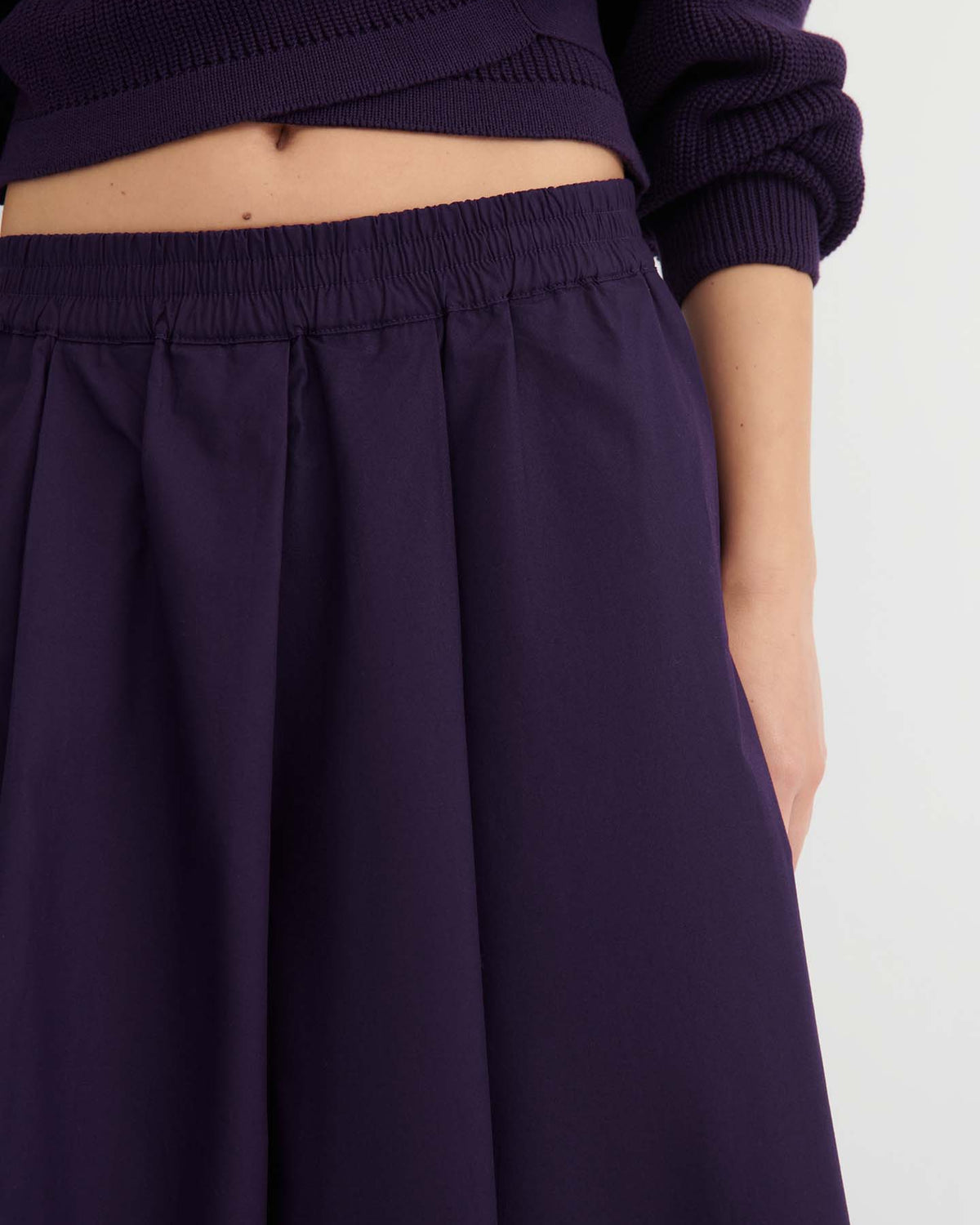 A person wearing the “Culottes – Dahlia,” which feature a dark purple pleated design with a natural waist, paired with a matching relaxed fit sweater, and one arm resting by their side.