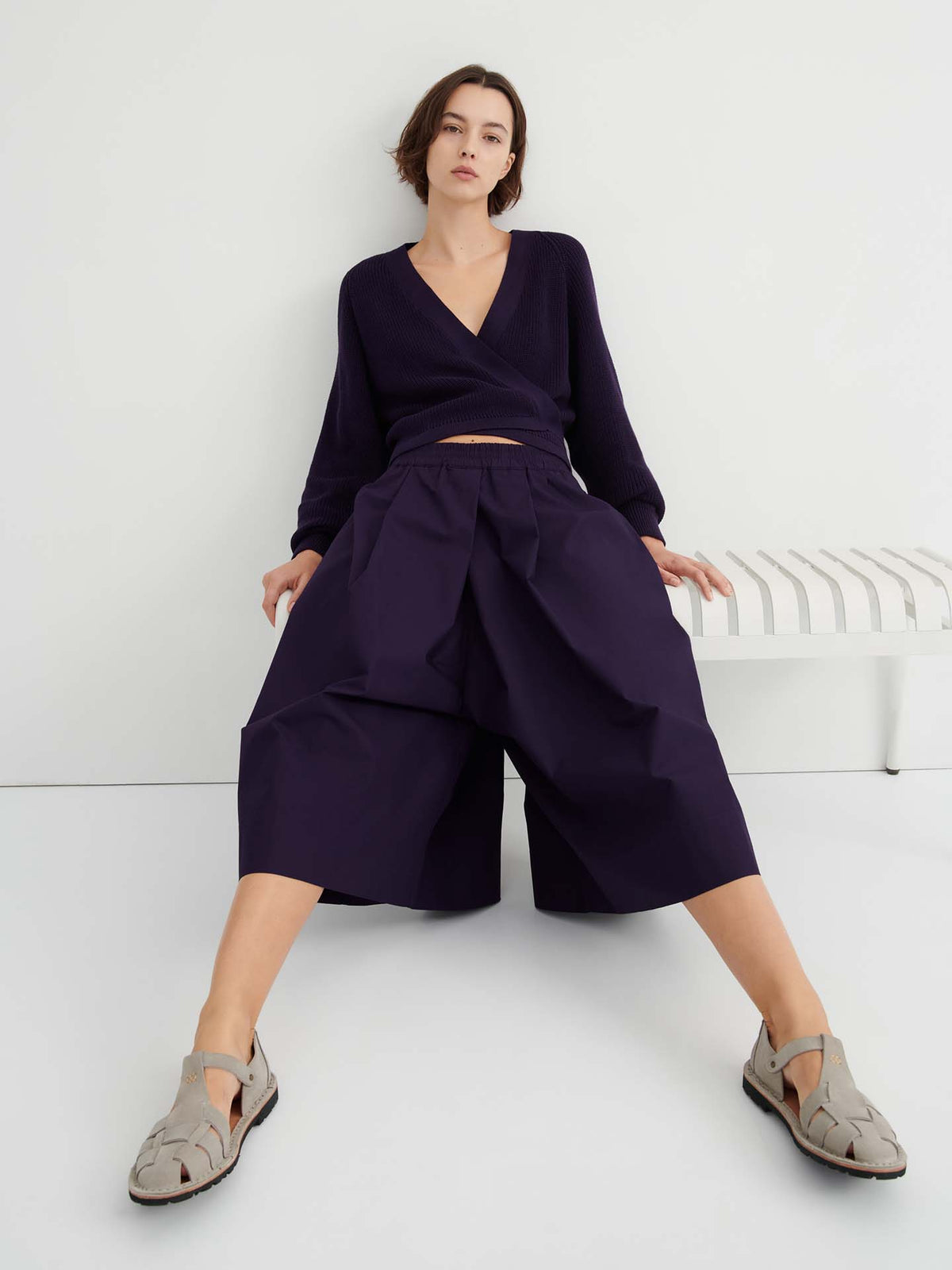 A person sits with legs apart on a white bench against a plain background, wearing a dark wrap top paired with the Dahlia wide, relaxed fit culottes featuring a natural waist.