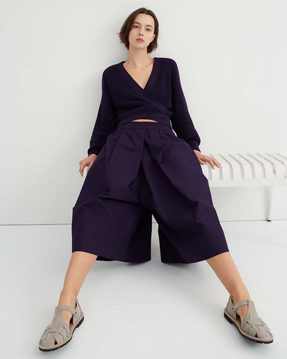 A person sits with legs apart on a white bench against a plain background, wearing a dark wrap top paired with the Dahlia wide, relaxed fit culottes featuring a natural waist.