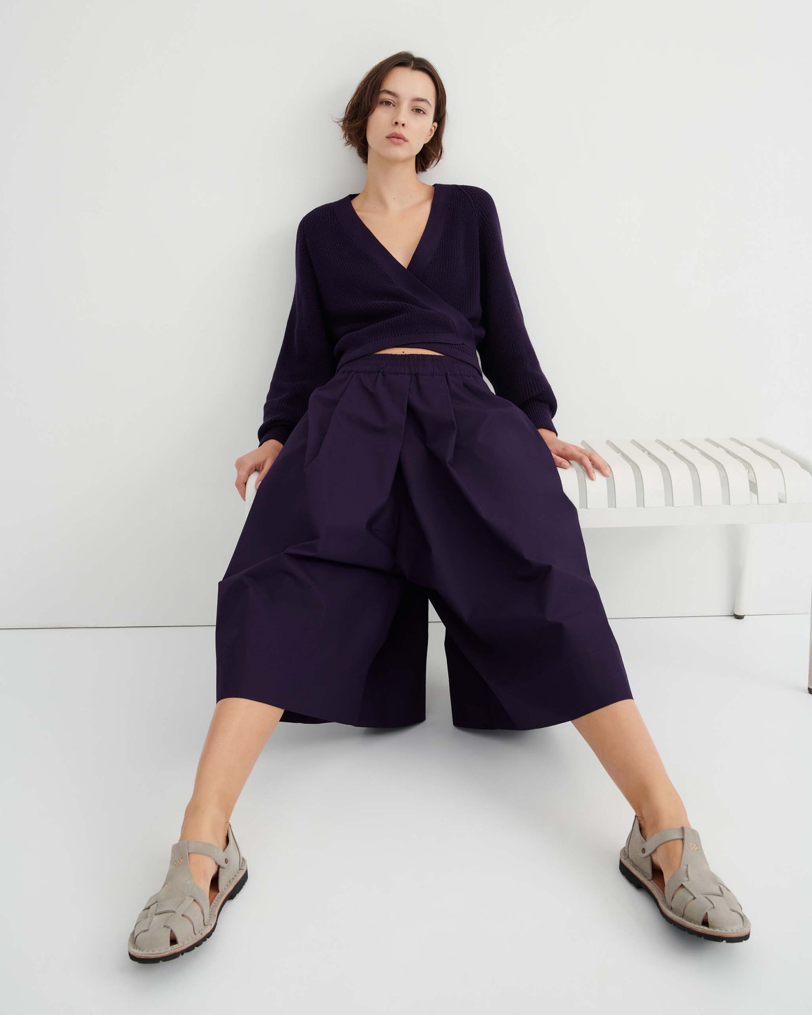 A person stands against a white wall wearing a dark purple wrap sweater, the Dahlia culottes—relaxed fit and sitting at the natural waist—with hands in their pockets, paired with gray sandals.