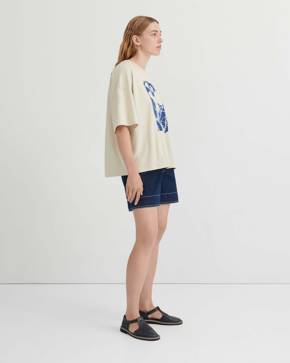 Person standing sideways against a white background, wearing a cream graphic t-shirt, dark blue shorts, and black slip-on shoes.
