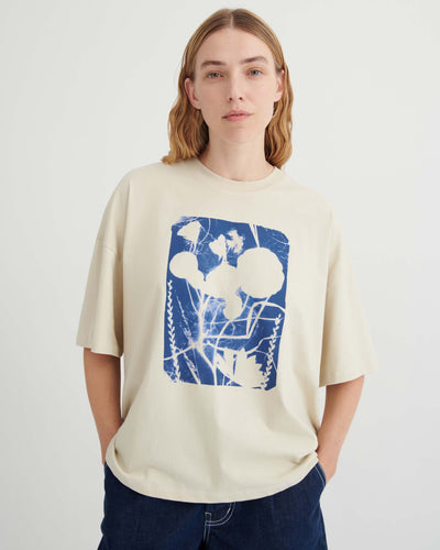 Person wearing a cream-colored T-shirt with a blue and white botanical graphic, standing against a plain light background, hands in pockets, looking at the camera.
