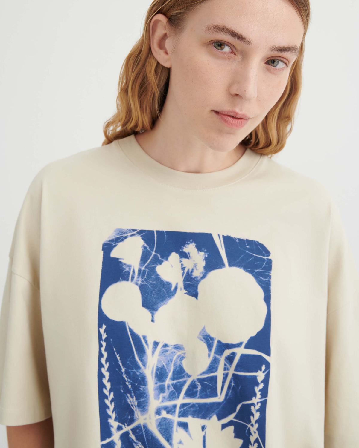 A person with light brown hair wears a cream-colored shirt featuring a blue and white botanical print, standing against a plain light background.