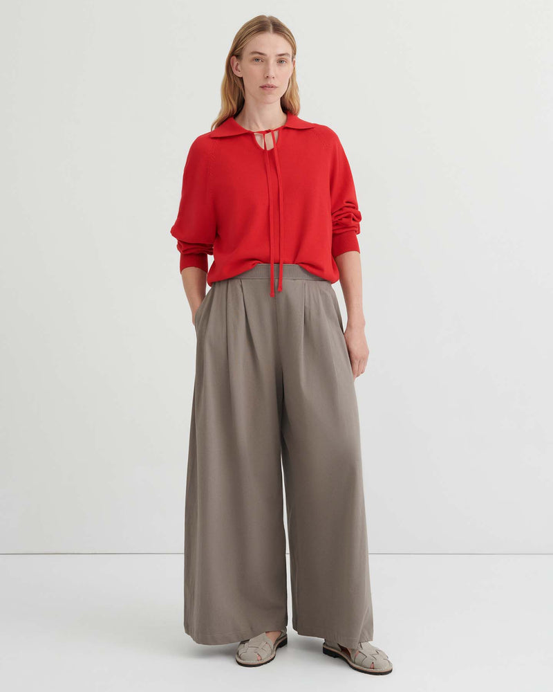 A person stands against a plain background wearing a red long-sleeve top with a tie neckline, the Drape Pant – Flint featuring an elastic waistband and wide leg in taupe, and beige shoes.