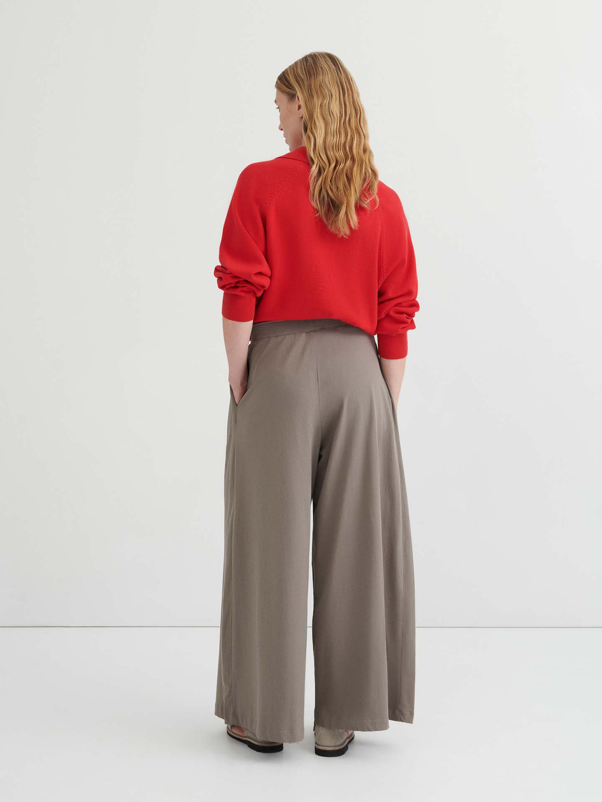 A person with long, wavy hair stands facing away, wearing a red sweater and the Drape Pant – Flint with an elastic waistband and wide-leg silhouette, hands in pockets, against a plain white background.