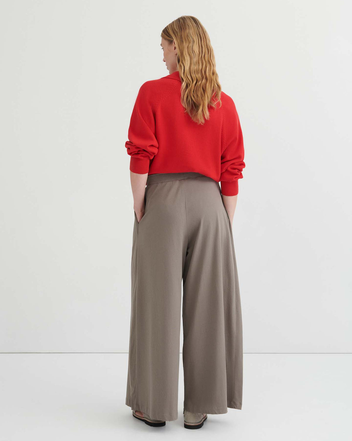 A person with long, wavy hair stands facing away, wearing a red sweater and the Drape Pant – Flint with an elastic waistband and wide-leg silhouette, hands in pockets, against a plain white background.