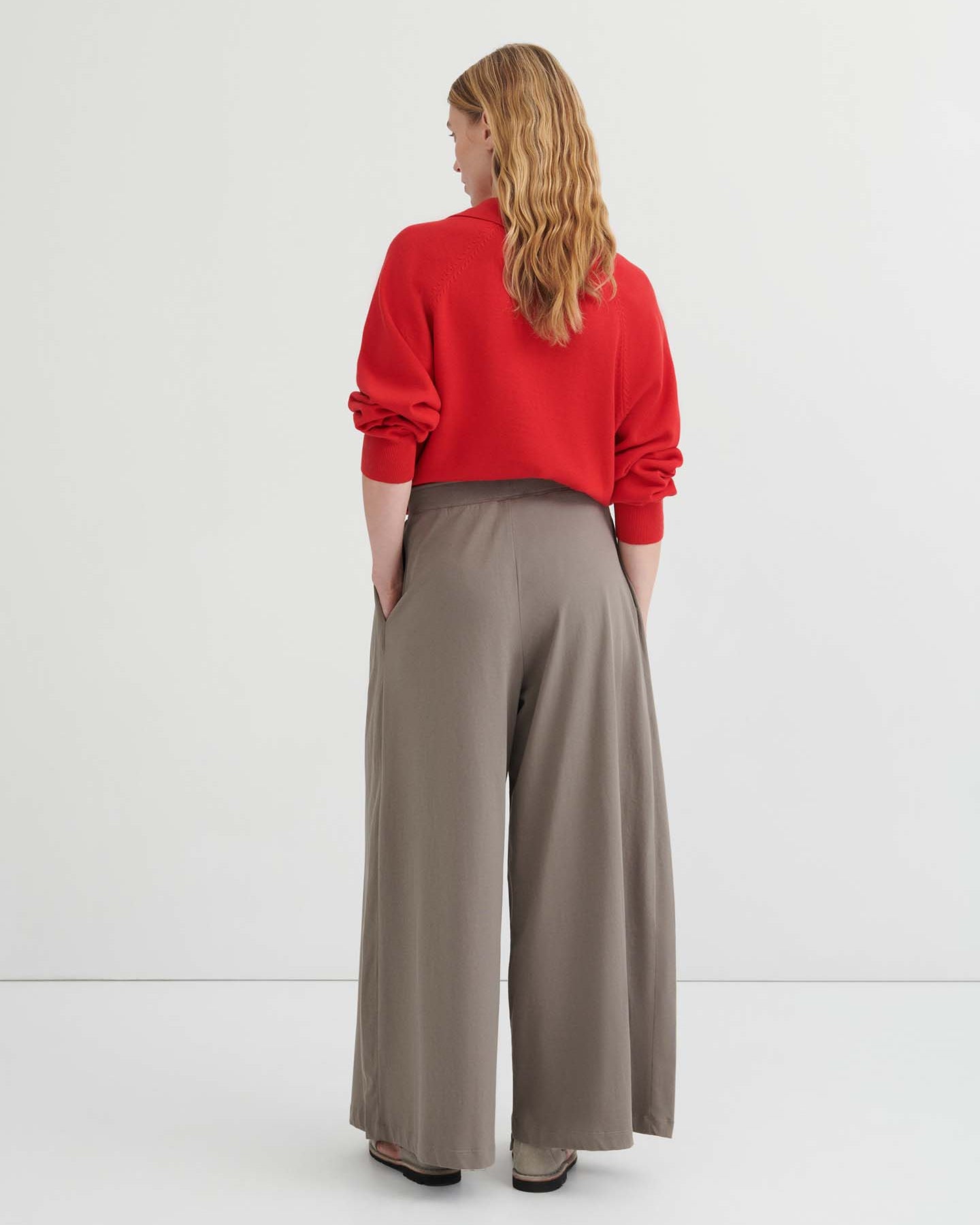 A person stands against a plain background wearing a red long-sleeve top with a tie neckline, the Drape Pant – Flint featuring an elastic waistband and wide leg in taupe, and beige shoes.