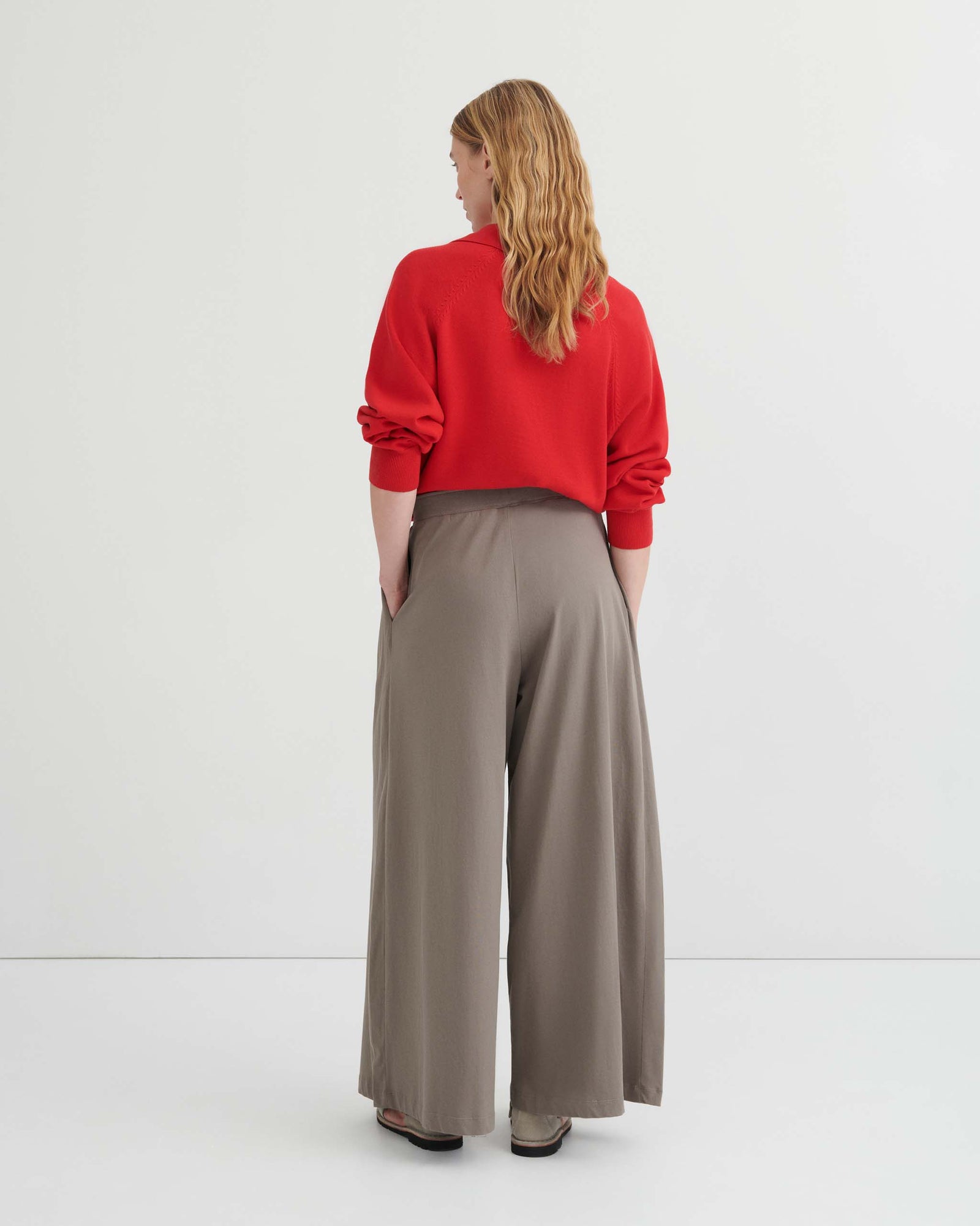 A person stands against a plain background wearing a red long-sleeve top with a tie neckline, the Drape Pant – Flint featuring an elastic waistband and wide leg in taupe, and beige shoes.