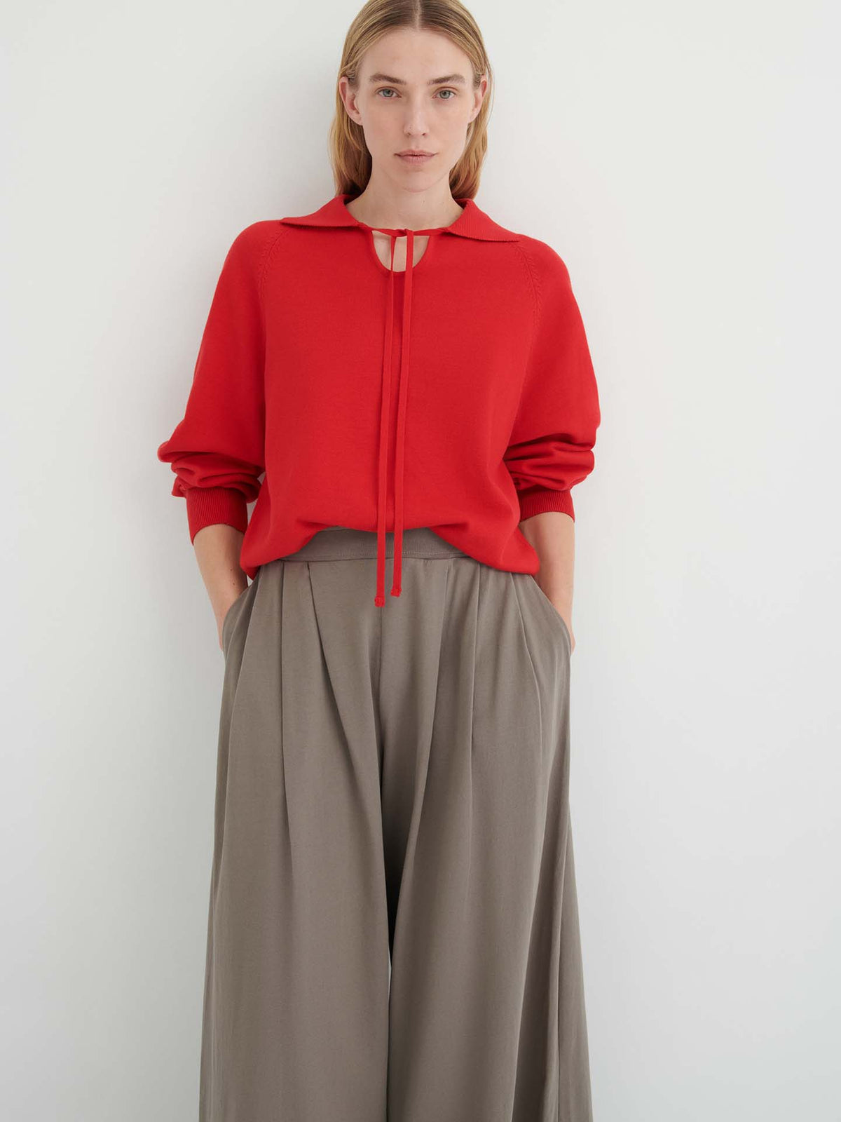 Person standing against a white wall, wearing a red long-sleeve top with tie detail and the Drape Pant – Flint, a high-waisted style with wide legs, elastic waistband, and hands in pockets.