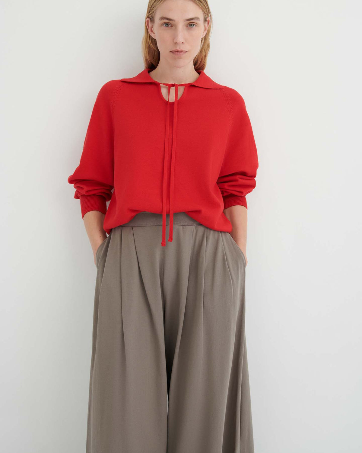 Person standing against a white wall, wearing a red long-sleeve top with tie detail and the Drape Pant – Flint, a high-waisted style with wide legs, elastic waistband, and hands in pockets.