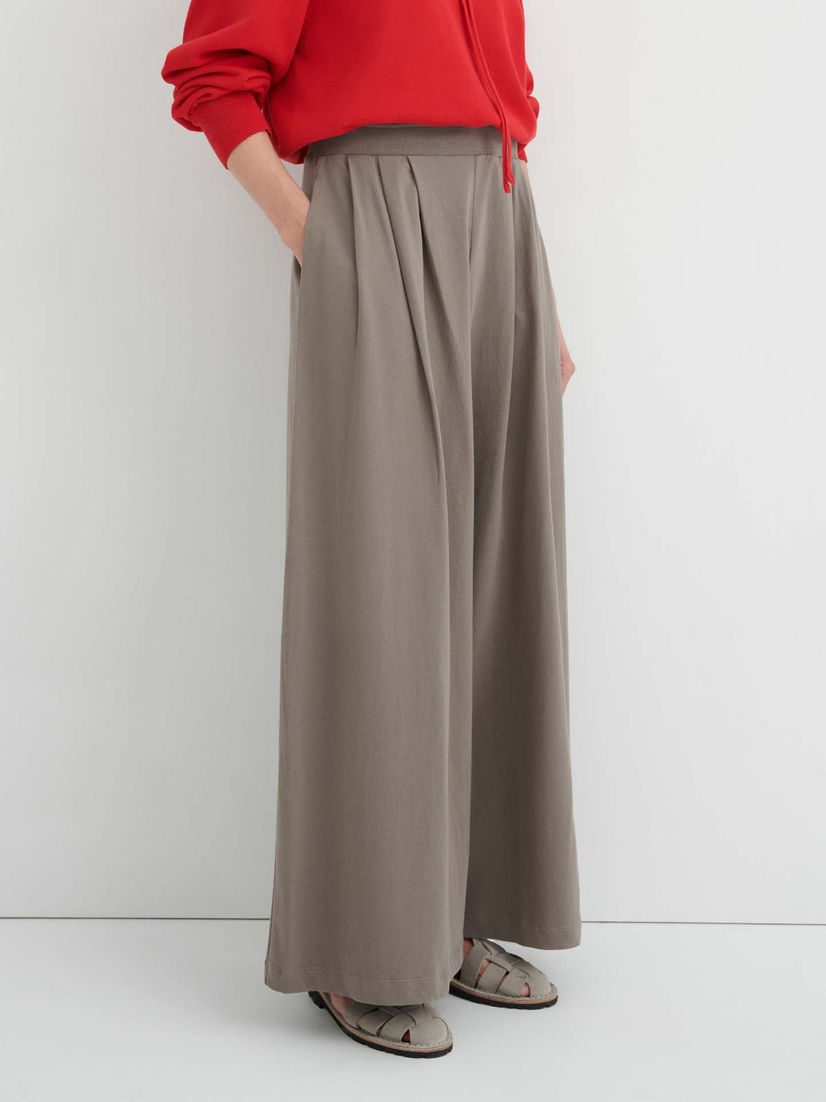 Person wearing a red long-sleeve top, the Drape Pant – Flint featuring a wide-leg silhouette and elastic waistband, paired with open-toed sandals, standing against a plain light-colored wall.