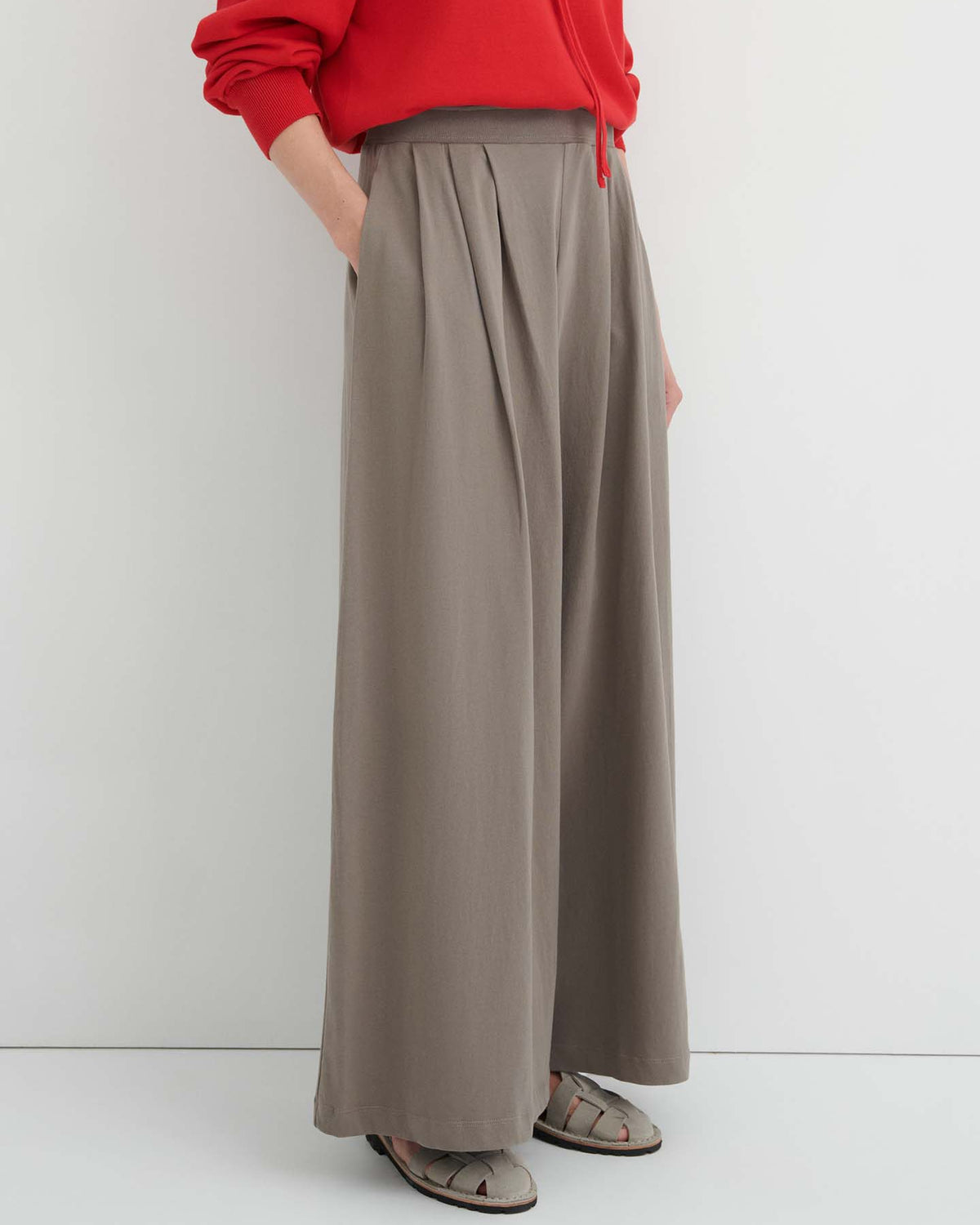 Person wearing a red long-sleeve top, the Drape Pant – Flint featuring a wide-leg silhouette and elastic waistband, paired with open-toed sandals, standing against a plain light-colored wall.