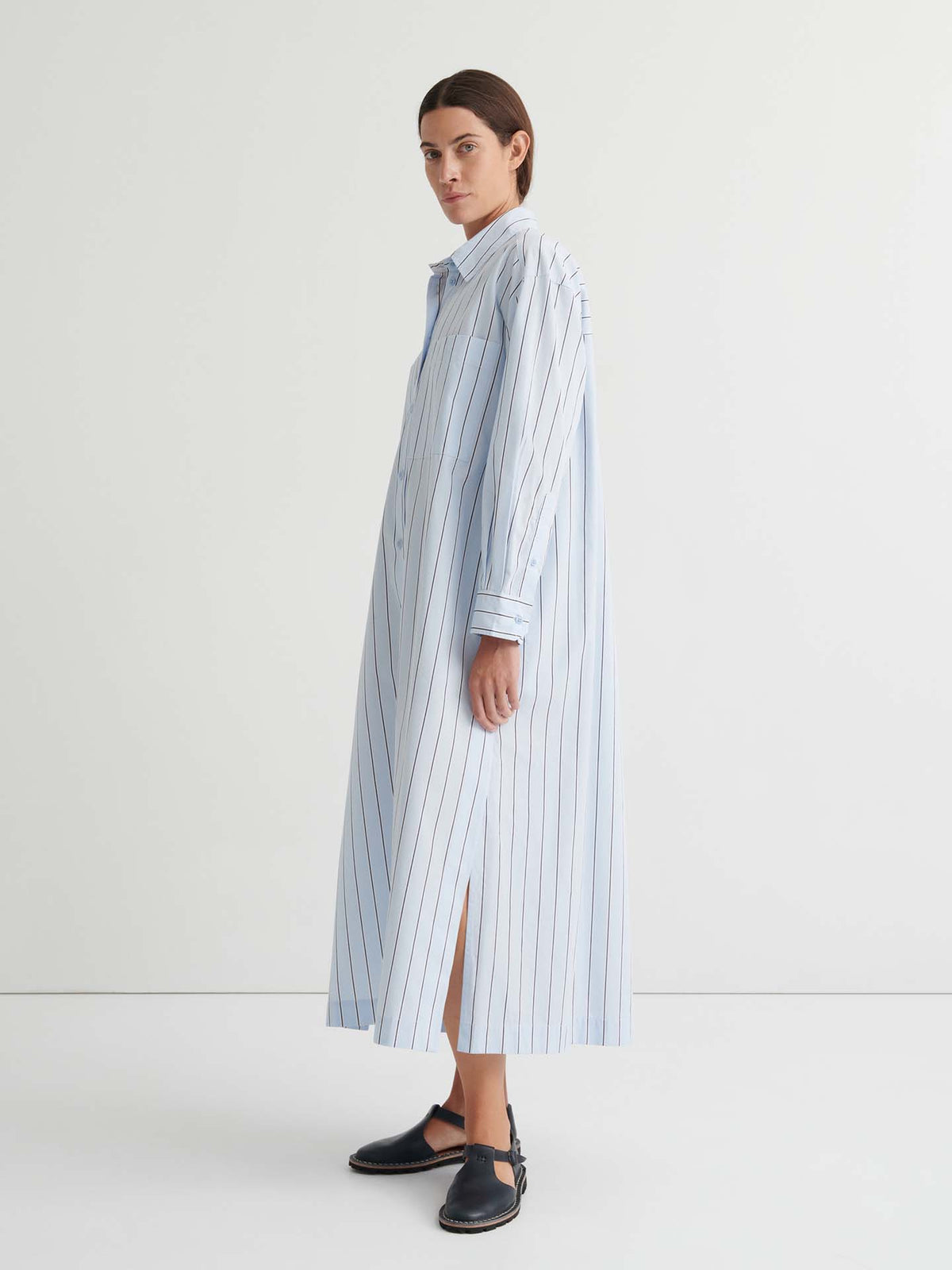 A person wearing the Elementary Dress – Cloud Stripe, an oversized, long, loose-fitting shirt dress with light blue and white vertical stripes and long sleeves, paired with black sandals, stands against a plain white background.
